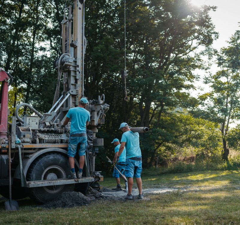 Water well drilling rig, truck preparing to boring dowin into the earth. Professional workers operating drilling machine, shoveling rubble. A rotary drill rig using bits to bore into ground and loosing the soil and rocks. Mud and muddy water is spitting out when drilling borewell. Banner with copy space.